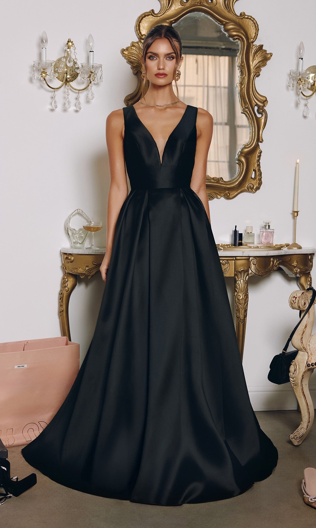 A woman stands indoors in front of a mirror and vanity table, wearing the Amoris T814 long black prom dress featuring a sleeveless mikado design, deep V-neckline, and floor-length A-line silhouette.